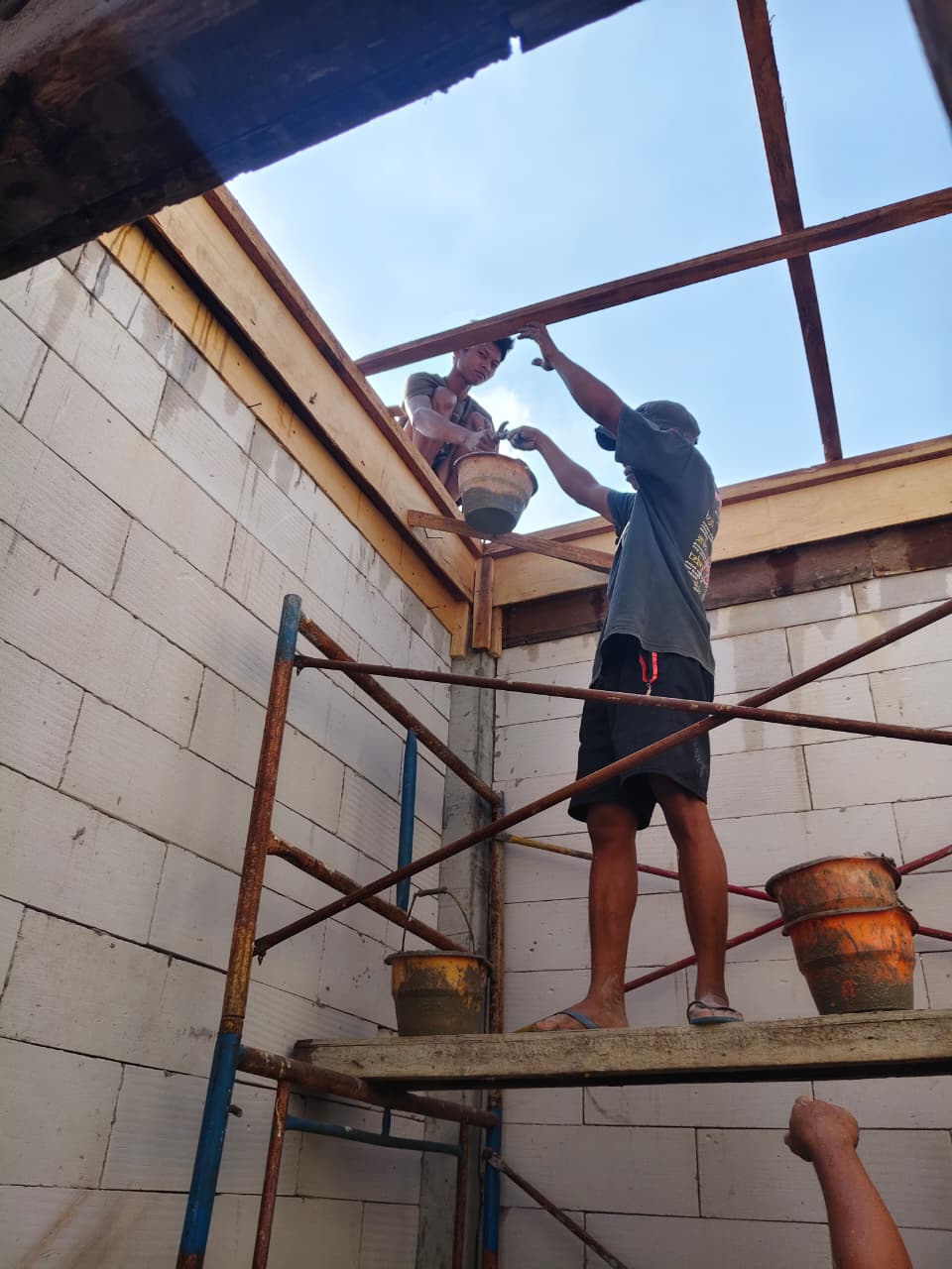 Pekerja menaikkan ember adukan beton ke balok reng atap menggunakan perancah